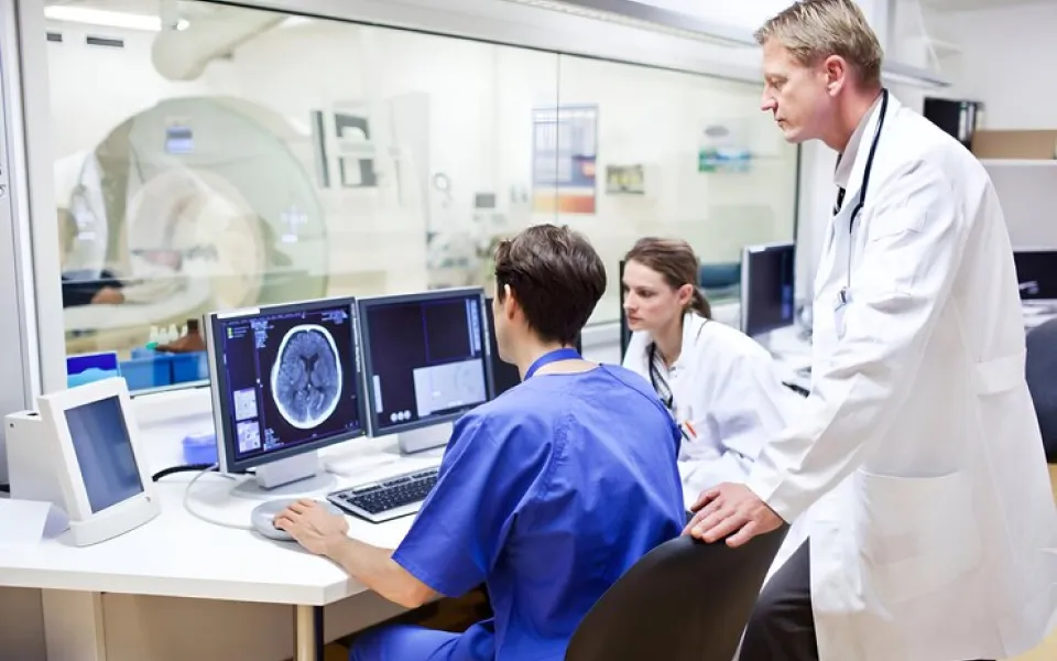 Senior doctor standing behind two doctors seated at a desk operating a PC displaying an MRI scan