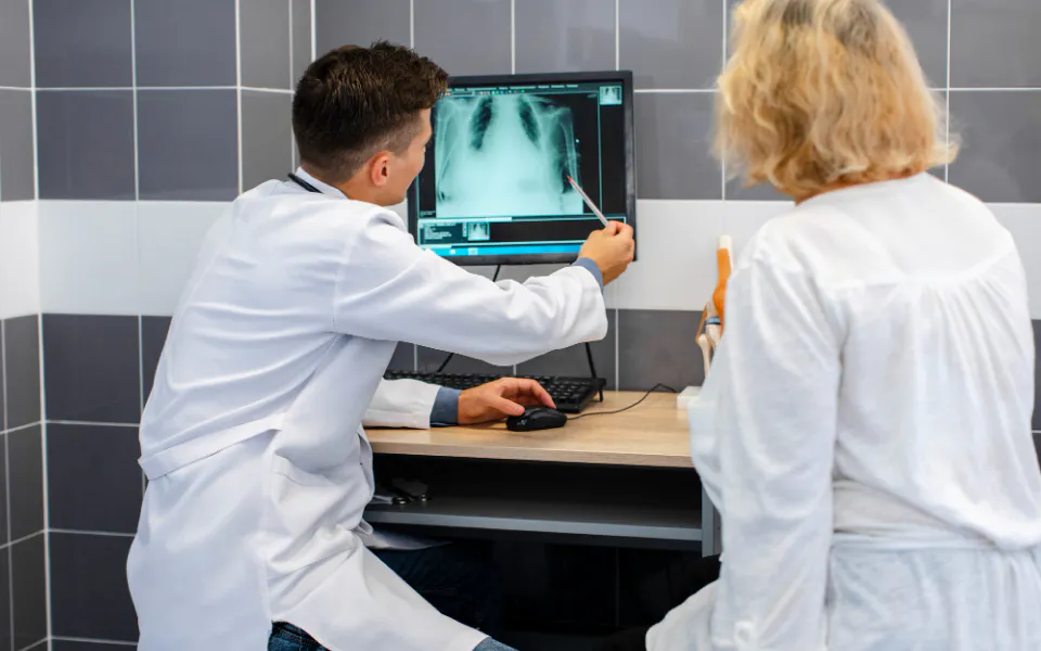 Doctor pointing at X-ray on monitor with pen, discussing findings with colleague using AI-assisted tools.