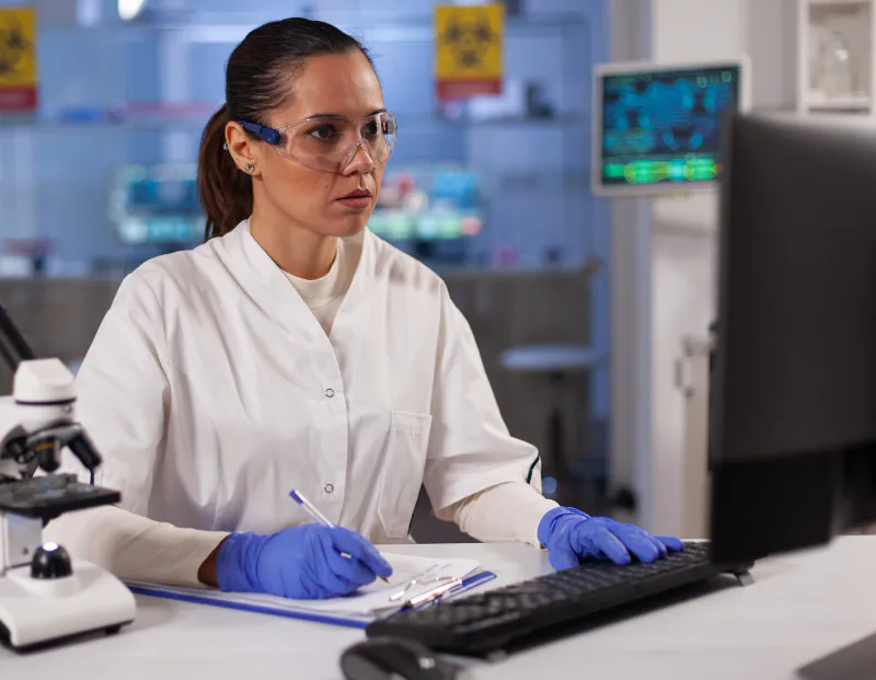 Female lab expert writing notes while reviewing advanced data on monitor screen.