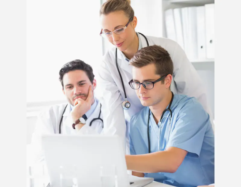 Senior doctor impressed, observing monitor with young doctor while female doctor operates PC.