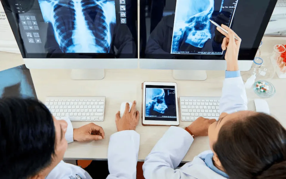 Two doctors sitting at a desk with one pointing at head scan reports displayed on a screen